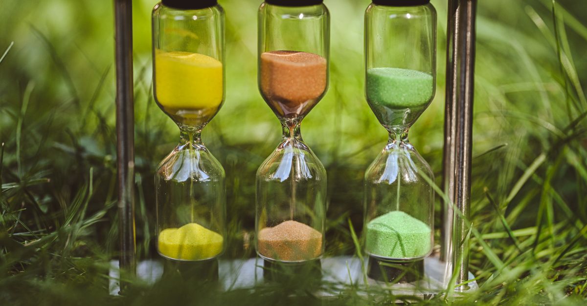 A vibrant triple hourglass with yellow, orange, and green sand set in a lush grassy meadow.