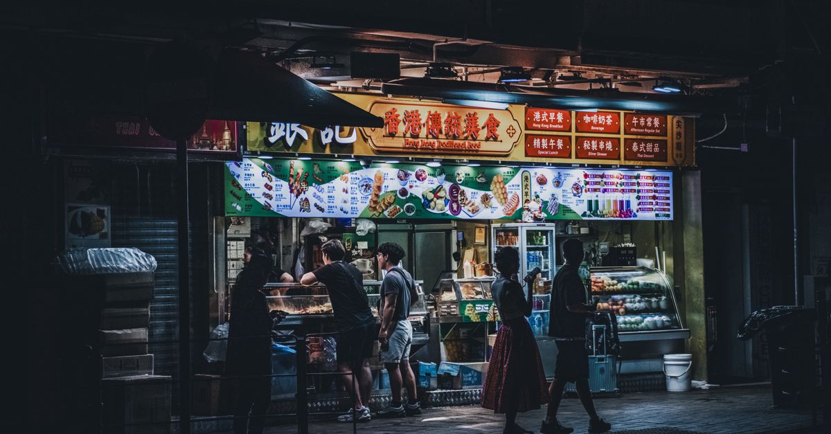 A bustling urban street food stall illuminated at night with passersby enjoying snacks.