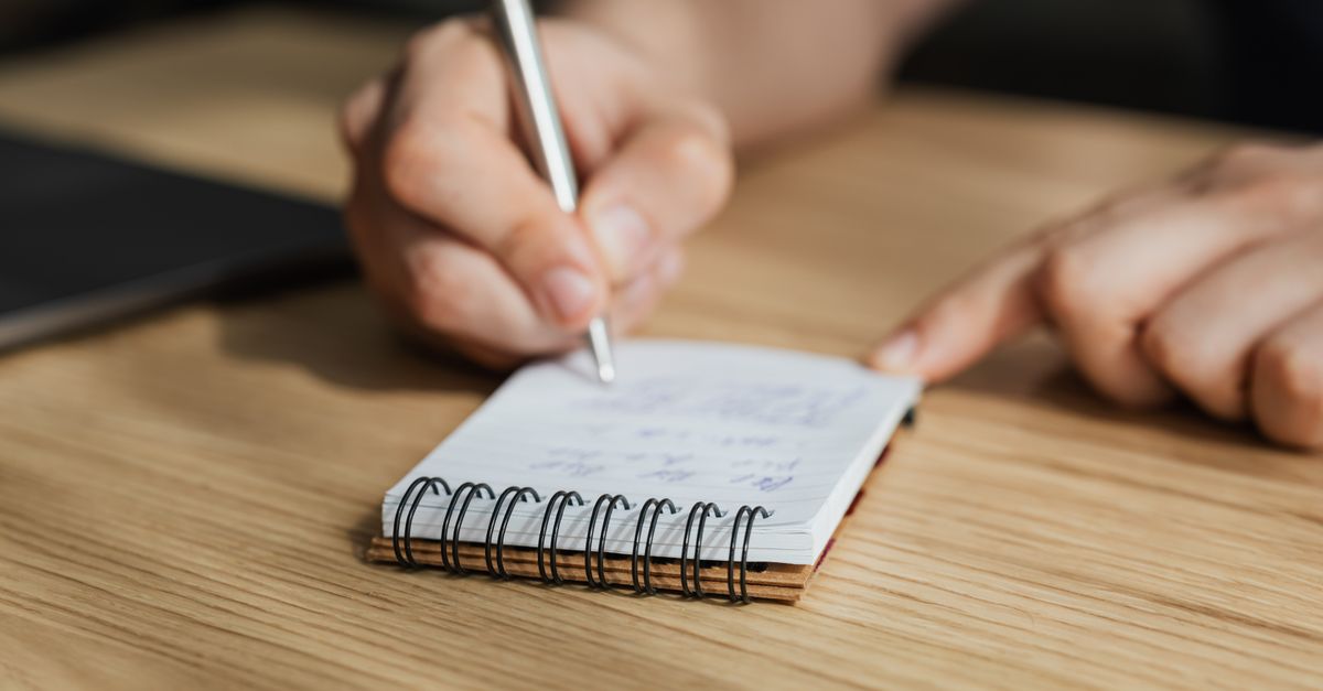 From above of crop faceless male student writing with pen in notebook at wooden desk during lesson