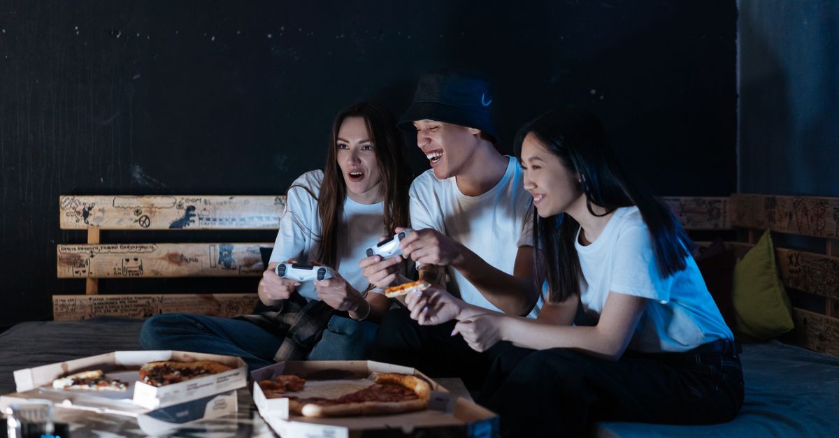 Group of diverse friends playing video games and eating pizza indoors, enjoying togetherness.
