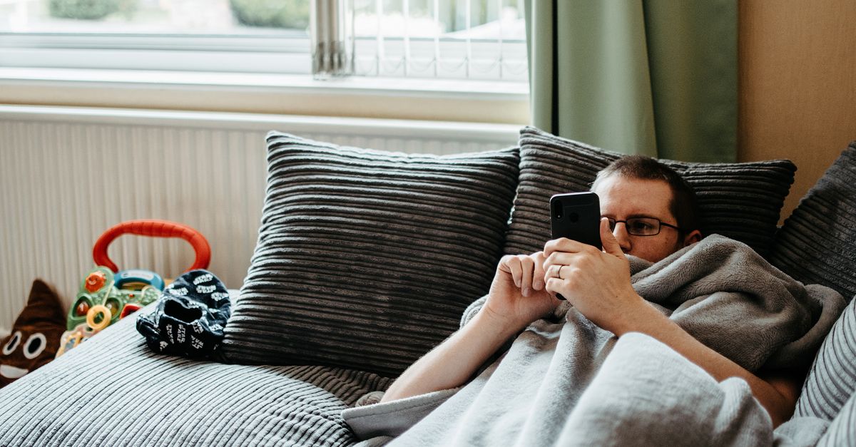 A man comfortably lying on a couch using a smartphone indoors, wrapped in a soft blanket.