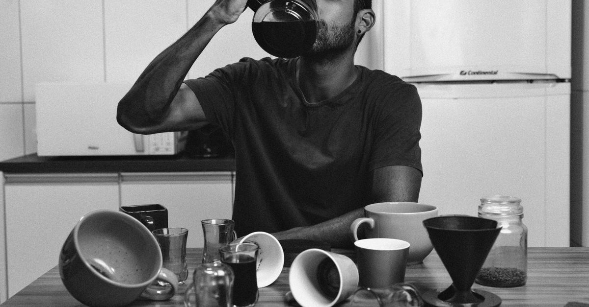 Man drinks coffee from a jug amid mugs and cups on a table.