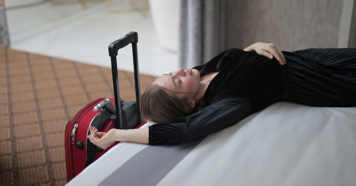 A woman resting on a hotel bed beside her suitcase, capturing travel fatigue.